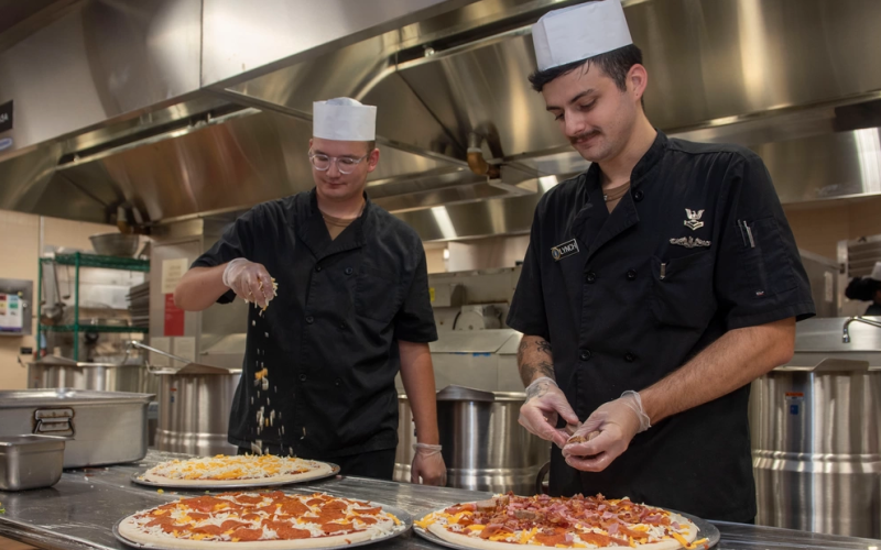 Culinary Specialists (Submarines) 2nd Class Simon Crews, left, and Culinary Specialist  Christian Lynch make pizza at the Pirates Cove Galley.